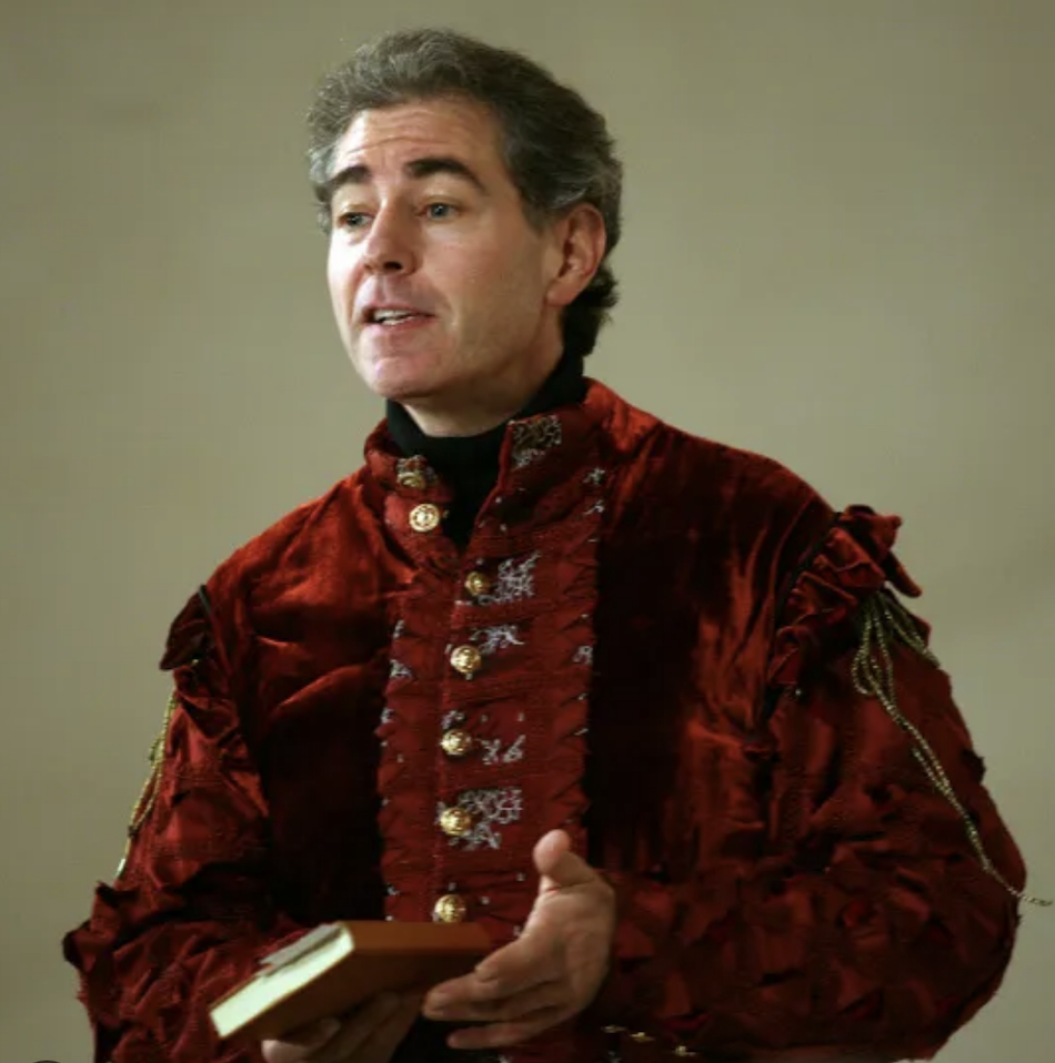 A man in a richly embroidered red velvet historical costume speaks animatedly, holding a small book.